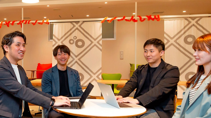 明るく装飾されたオフィスラウンジで、丸テーブルにノートPCを広げ笑顔で語り合う4人のメンバー。