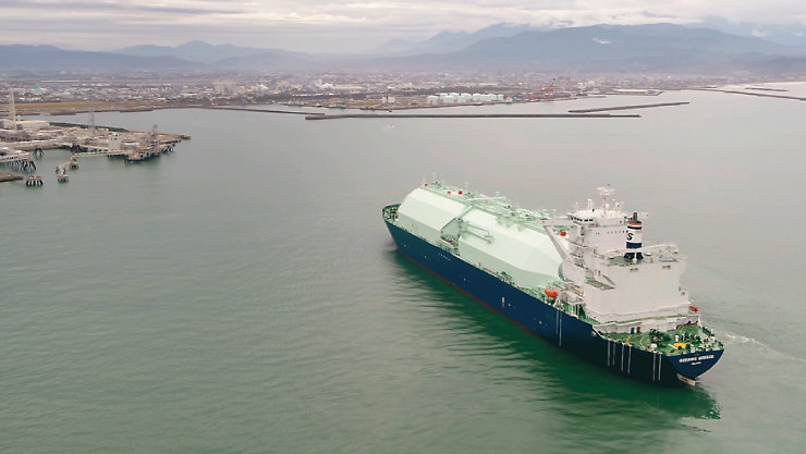 曇り空の下、工業地帯と山々を背景に湾内を航行するLNGタンカー。