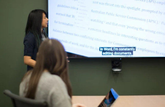 Eine Frau hält eine Präsentation, auf dem Bildschirm zu ihrer Seite sind Untertitel zu sehen