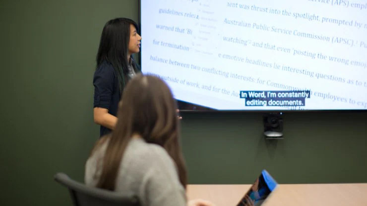 Eine Frau hält eine Präsentation, auf dem Bildschirm zu ihrer Seite sind Untertitel zu sehen