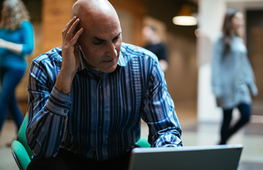 Ein Mann bedient sitzend einen Laptop und hält die Hand am rechten Ohr, hinter ihm laufen Personen vorbei