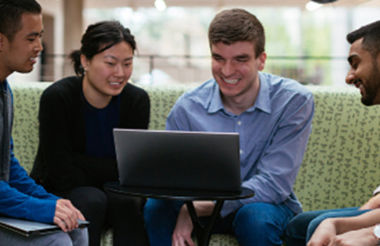 Eine Personengruppe sitzt um einen runden Tisch zusammen, auf dem ein Laptop steht und sieht lächelnd auf den Bildschirm