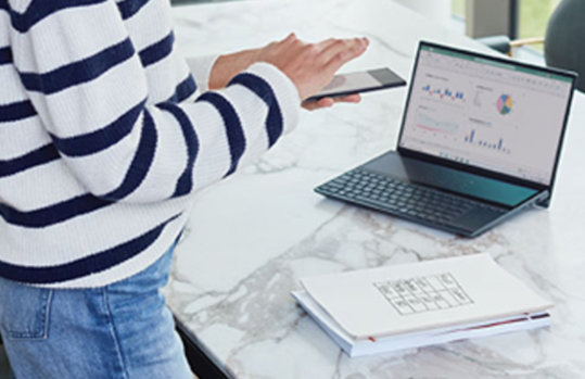 Eine Person bedient ein Smartphone, vor ihr befindet sich ein großer Marmortisch auf dem ein Laptop sowie Arbeitsunterlagen platziert sind