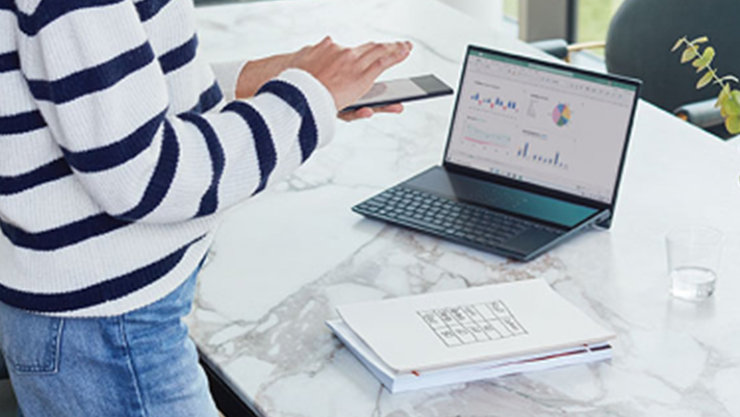 Eine Person bedient ein Smartphone, vor ihr befindet sich ein großer Marmortisch auf dem ein Laptop sowie Arbeitsunterlagen platziert sind