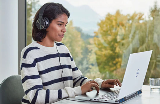 Eine Frau sitzt neben einer Glaswand und bedient einen Laptop, sie trägt geräuschreduzierende Kopfhörer
