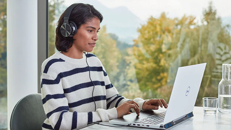 Eine Frau sitzt neben einer Glaswand und bedient einen Laptop, sie trägt geräuschreduzierende Kopfhörer