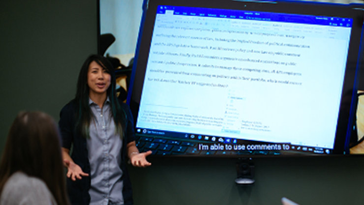 Eine Frau spricht, auf dem Bildschirm neben ihr wird ein Video mit Untertiteln angezeigt