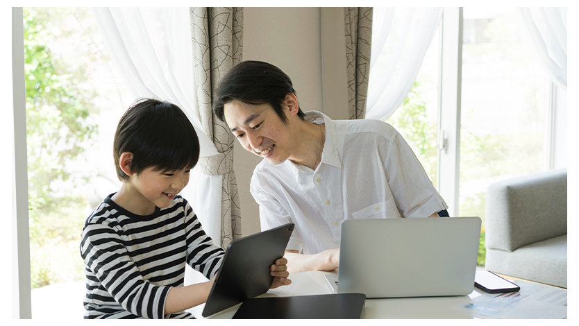 ノート PC での作業をしながら子どもが操作するタブレットをのぞき込む親子