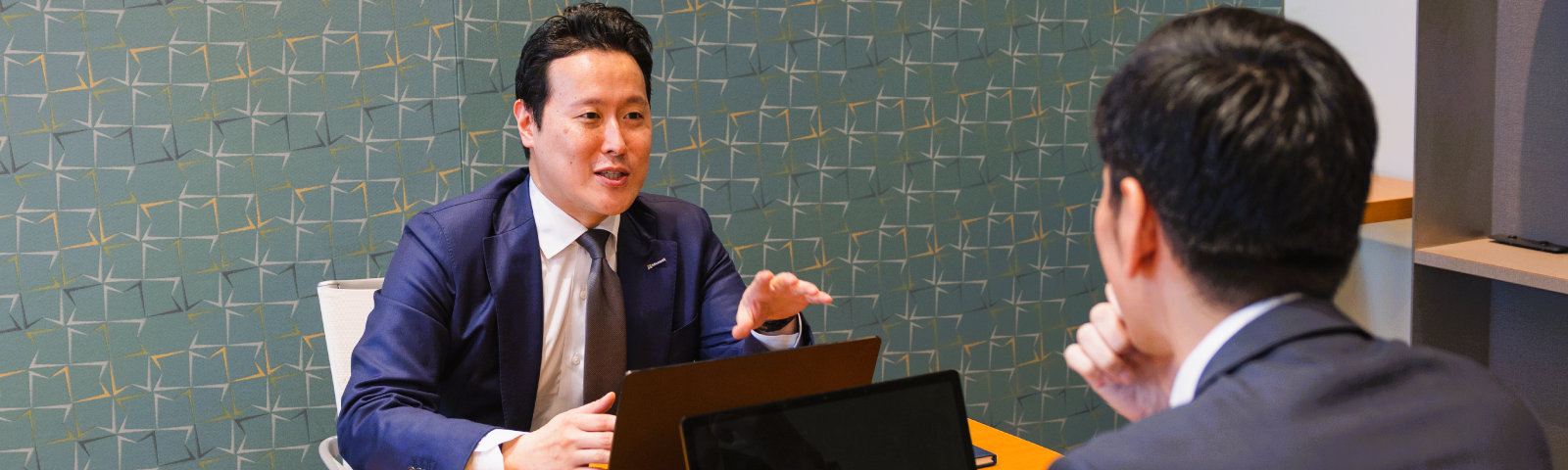 緑色のユニークなパターンの壁紙の社内で他の社員と話している様子のShota