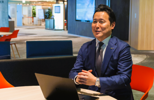 オレンジ色の椅子に座り、パソコンを前に置いて話しているShota