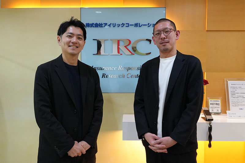 株式会社アイリックコーポレーション の受付エリアで、IRCロゴの前に立つ2名のビジネスパーソン