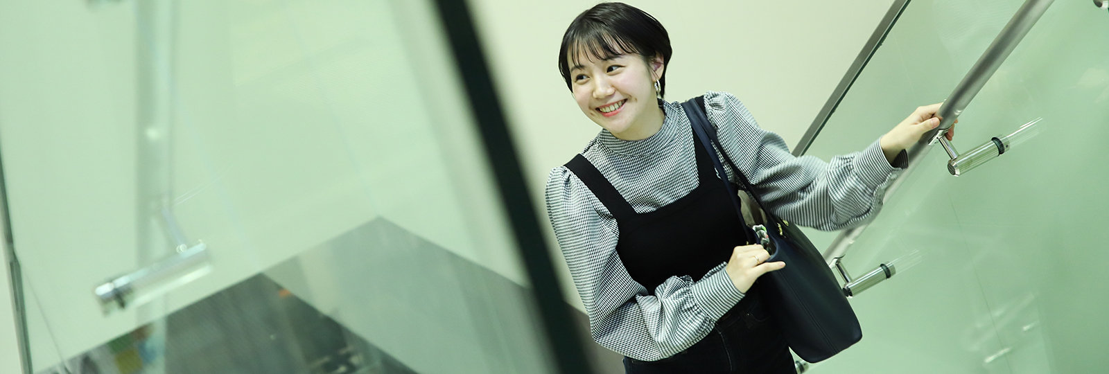 女性は片手で手すりを持ち、もう一方の手でバッグを持ち、ガラスの階段を微笑みながら上っています。