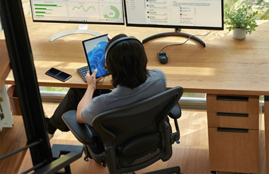 ヘッドフォンを装着し、2 台の大型 PC の前の椅子に座って手持ちのタブレット画面を見ている男性