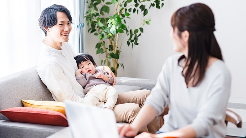 自宅でリモートワークをする母親とソファで子どもを抱く父親