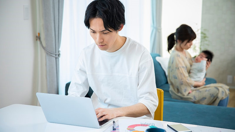 自宅のテーブルで仕事をする父親と後ろで子どもを抱きかかえる母親