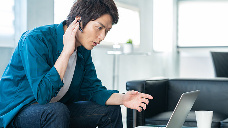 ソファに座って ノート PC を見ながら携帯電話で通話をする人物