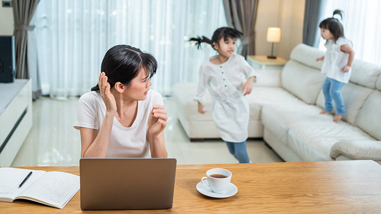 自宅のテーブルで仕事をしている女性と、後ろのソファで遊ぶ子供たち
