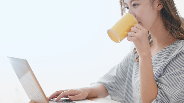 カップで飲み物を飲みながらノート PC を操作する人物