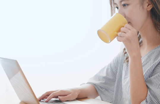 カップで飲み物を飲みながらノート PC を操作する人物