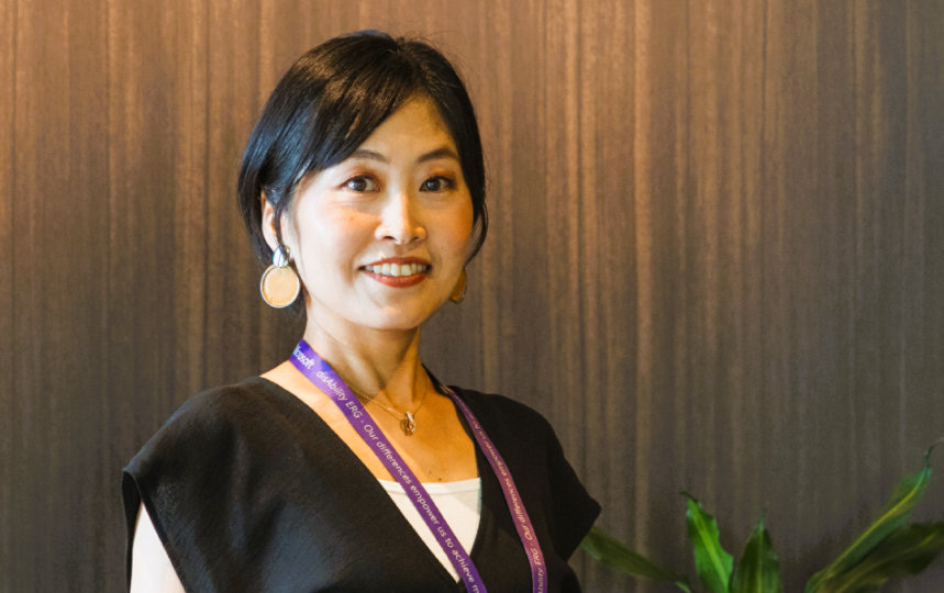 観葉植物に囲まれたMadokaが、木目調の壁の前に立っている室内風景