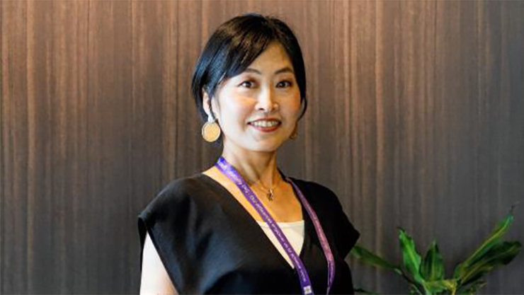 観葉植物に囲まれたMadokaが、木目調の壁の前に立っている室内風景