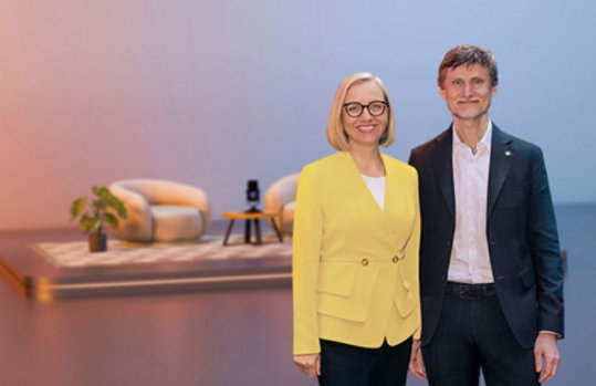 Zwei Personen in Business-Kleidung vor einem stilisierten Hintergrund mit Möbeln.