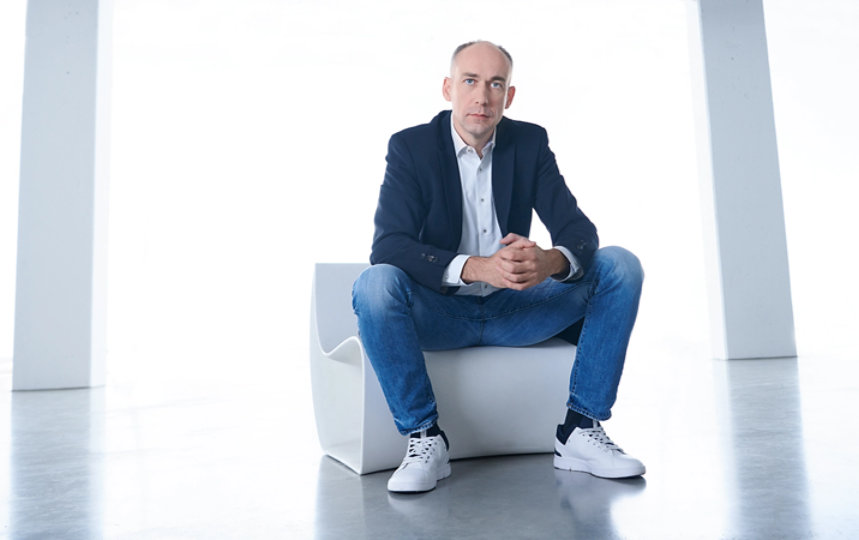Matthias Gohl mit Jeans, weißem Hemd und blauen Jackett sitzt auf einem weißen Stuhl.