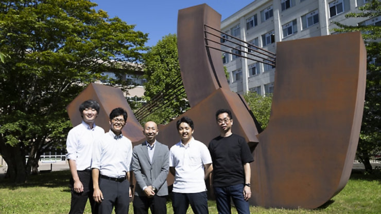 緑の木々と建物を背景に、大学キャンパスの大きな抽象的な金属彫刻の前に並ぶ5人の男性。