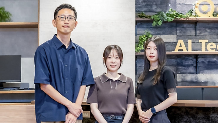 木製の棚や植物、「AI Te」の文字が見えるモダンなオフィスに立つ3人のプロフェッショナル。