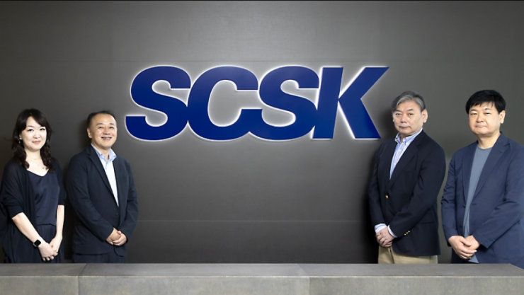 SCSKの企業ロゴの前に立つ4人のプロフェッショナル。