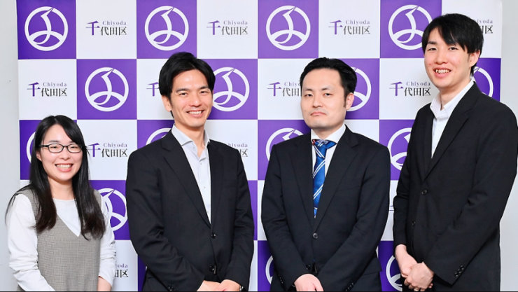 千代田区のロゴ入りバックドロップの前に立ち、カメラに向かって笑顔の4人のプロフェッショナル。