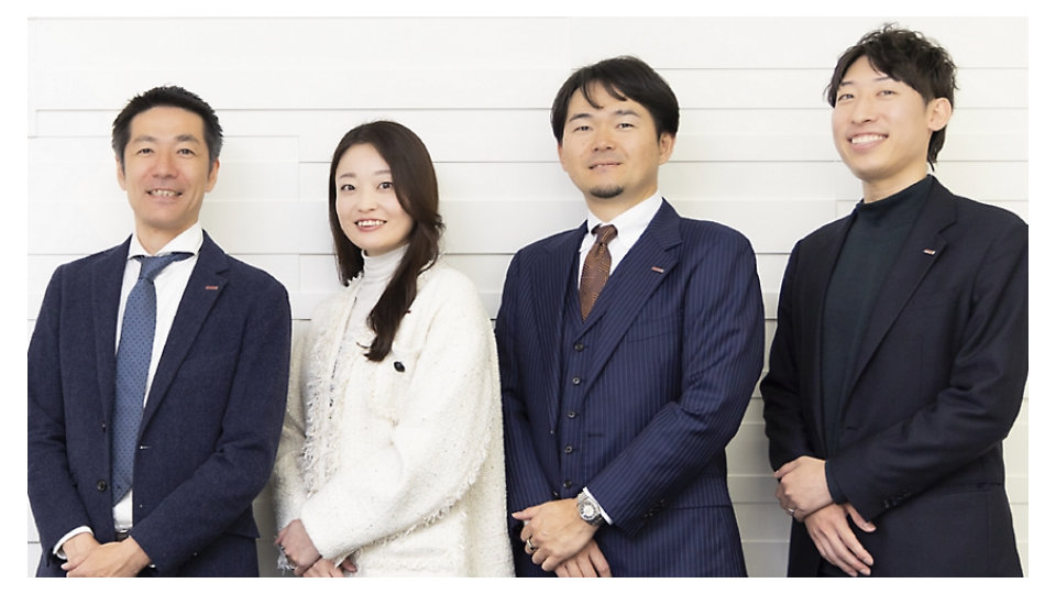 白い壁の前に並んで立つ、フォーマルな服装の笑顔の日本人ビジネスプロフェッショナル4名。