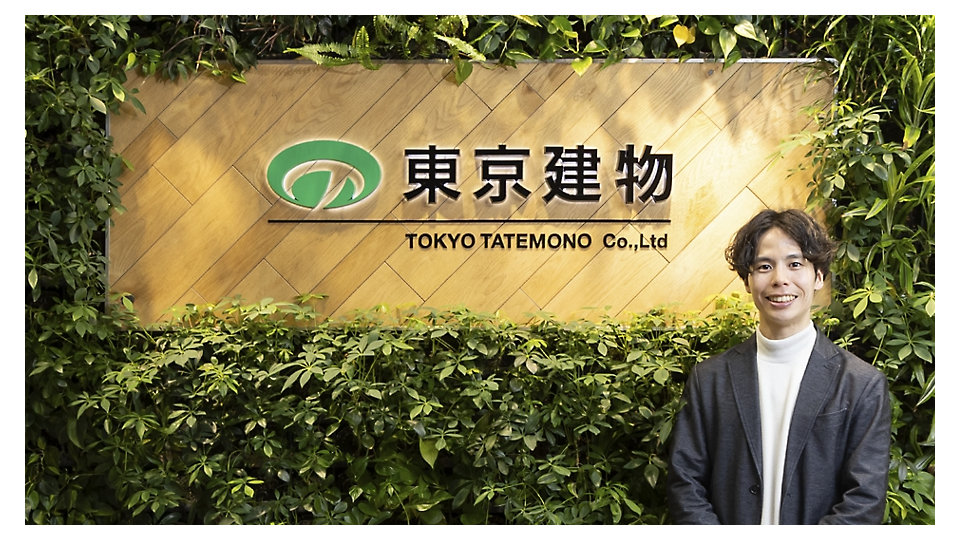 緑の植物に囲まれた東京建物（TOKYO TATEMONO Co., Ltd）の企業看板の前に立つグレーのブレザーを着た笑顔の男性。