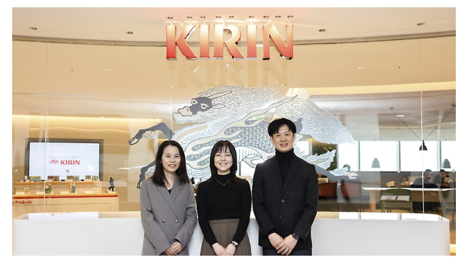KIRINの企業ロゴと麒麟の壁画を背景に、オフィスロビーで笑顔の3人のプロフェッショナル。