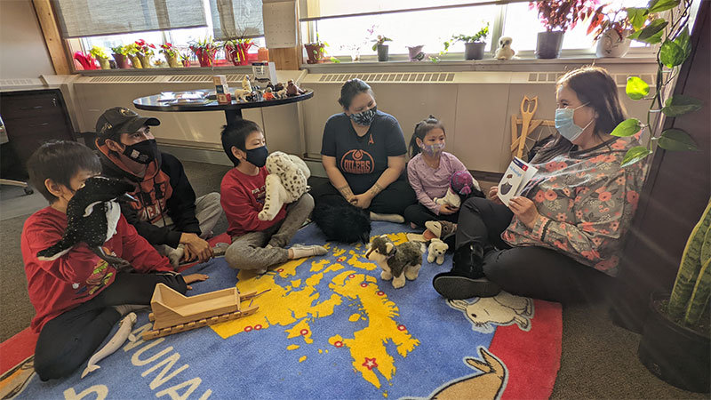 L’honorable ministre Pamela Hakongak Gross lire un livre d’images à un groupe d’enfants tenant des animaux en peluche et assis sur le sol