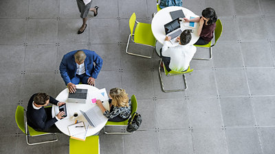 2 つの丸いテーブルでそれぞれノート PC で作業をする 5 人の人物