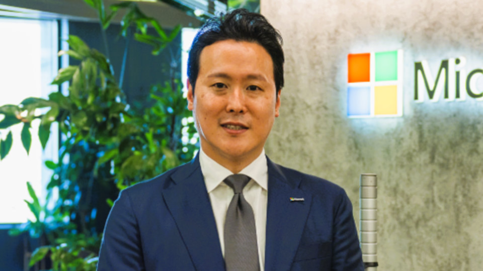 Microsoftのロゴが後ろに見える社内で立っているShota