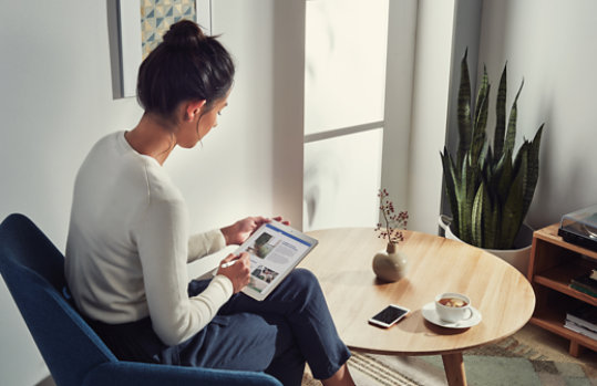 屋内で一人の女性がタブレットを見ている。テーブルの上には一杯のコーヒー、スマートフォン、花瓶がある。