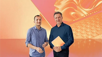 Zwei Männer stehen vor abstraktem, orangefarbenem Hintergrund, der einen Laptop darstellt. Die rechte Person hält einen Laptop in den Händen.