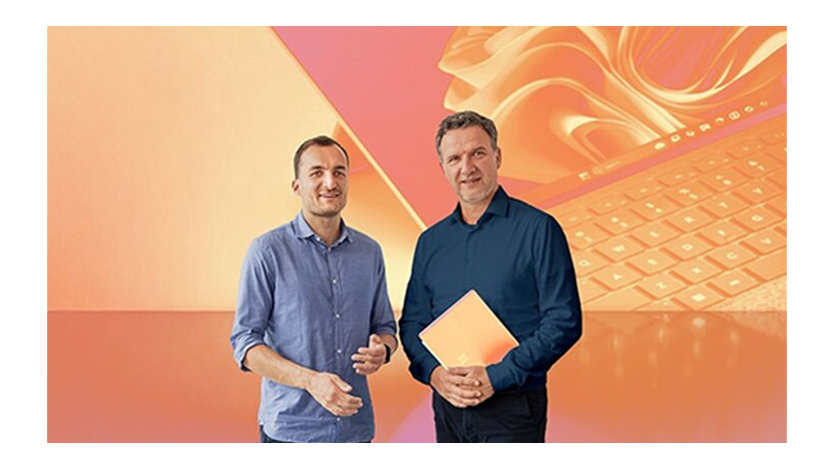 Zwei Männer stehen vor abstraktem, orangefarbenem Hintergrund, der einen Laptop darstellt. Die rechte Person hält einen Laptop in den Händen.