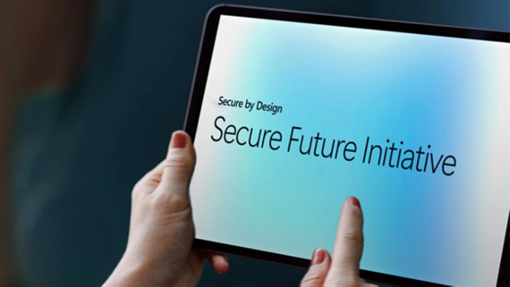 Eine Person hält ein Tablet mit der Aufschrift 'Secure by Design' und 'Secure Future Initiative'.