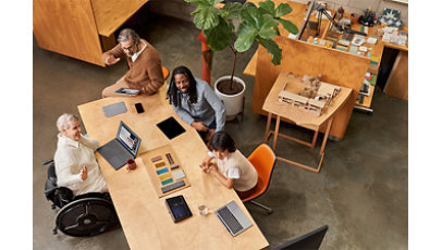 オフィスの上の方から見た写真：大きなテーブルに数人の男女が座り、楽しそうに話し合っている。机の上にはパソコンやタブレットなどが置いてあり、左側の女性は車いすに乗っている