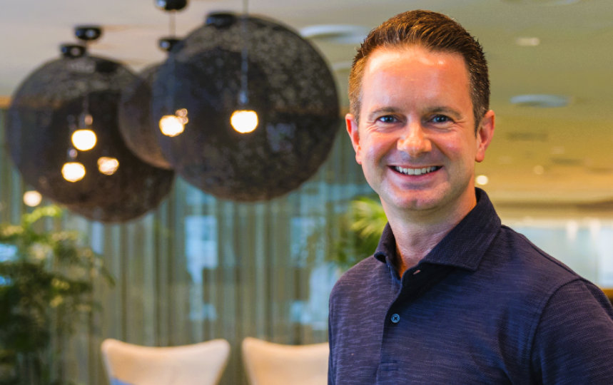 黒くて丸い照明器具が後ろに見えている室内で笑顔で立っているJason