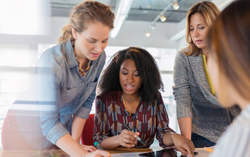 Vier Frauen im Gespräch. Eine der Frauen sitzt am Tisch und hat einen Laptop vor sich stehen.