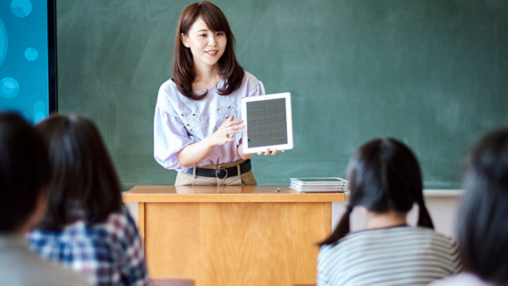 教室の黒板の前で、女性教師が教卓から生徒たちに向けて白いタブレットを掲げ、笑顔で説明を行っている場面。手前には授業を聞く生徒たちの後ろ姿があり、教師が持つ端末の画面には書類のようなグレーのモックテキストが配置されています。教卓の上には重ねられた数枚の端末が置かれています。