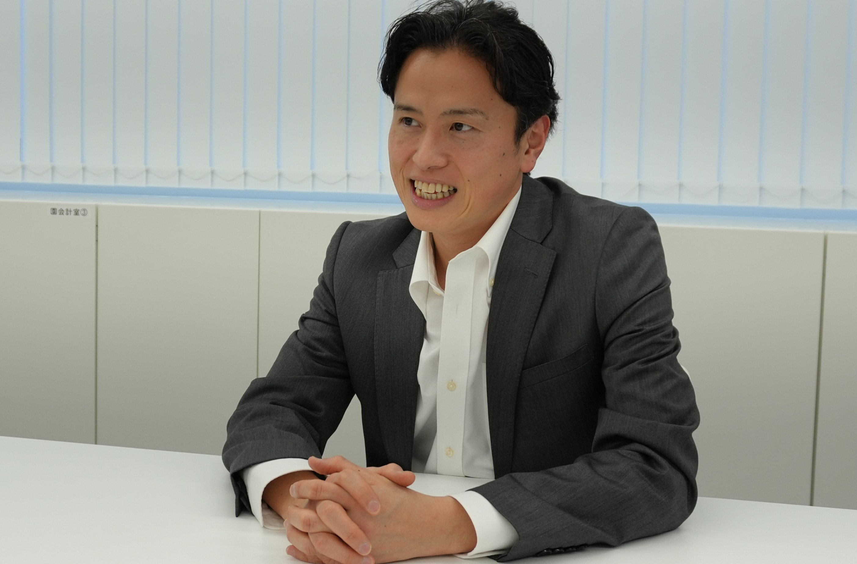 グレーのスーツと白いシャツを着た男性が白いテーブルに座り、手を組んで微笑んでいます。部屋には縦型のブラインドが見えます。