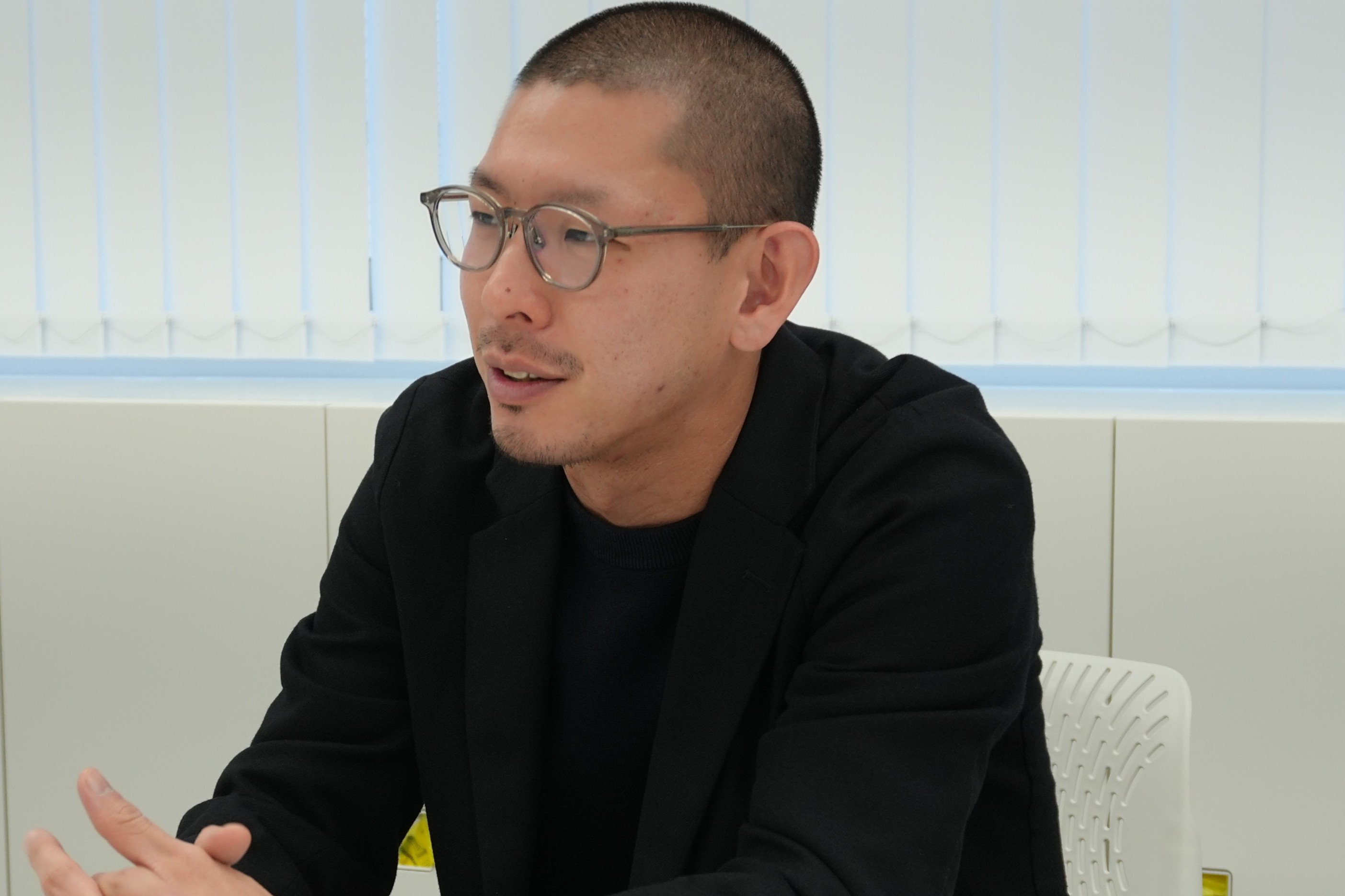 眼鏡をかけ、坊主頭で黒いブレザーを着た男性が、会議室でバーティカルブラインドを背景に話しながら身振り手振りしている様子。