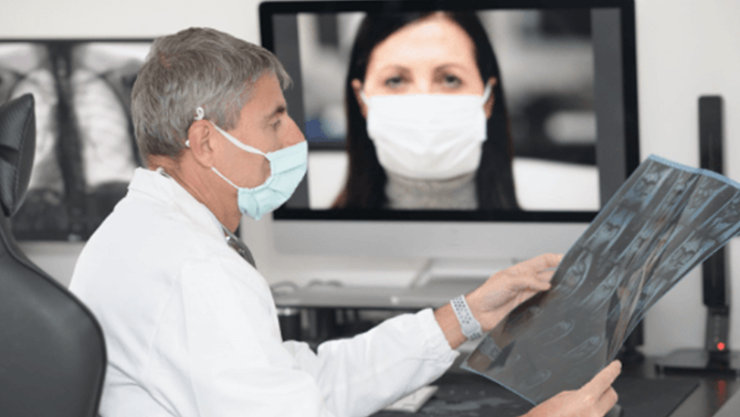 Lekarz z maseczce siedzi za biurkiem i ogląda zdjęcie rentgenowskie, w tle za nim stoi monitor, a na nim widać lekarkę w maseczce podczas spotkania na Teams