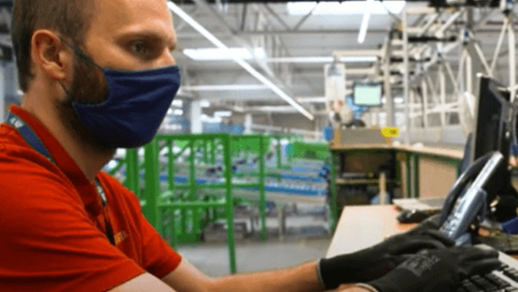 Pracownik w maseczce patrzy w monitor stojący na wysokim blacie, w tle hala produkcyjna