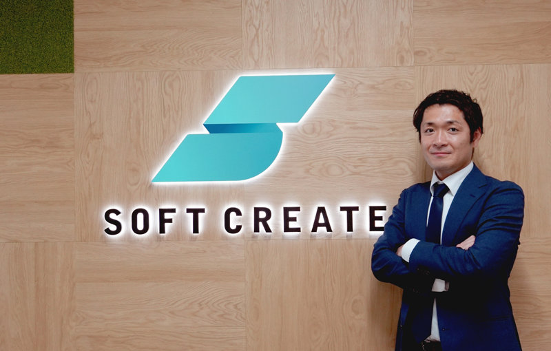 Soft Createの光るロゴが掲げられた木目調の壁の前に立ち、腕を組んでいるダークブルースーツ姿の男性。